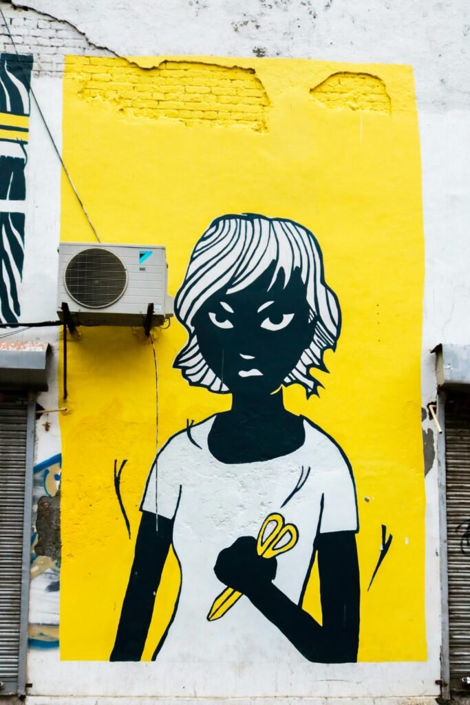 Colorful street art mural depicting a figure with scissors on a bright yellow wall in Delhi, India.